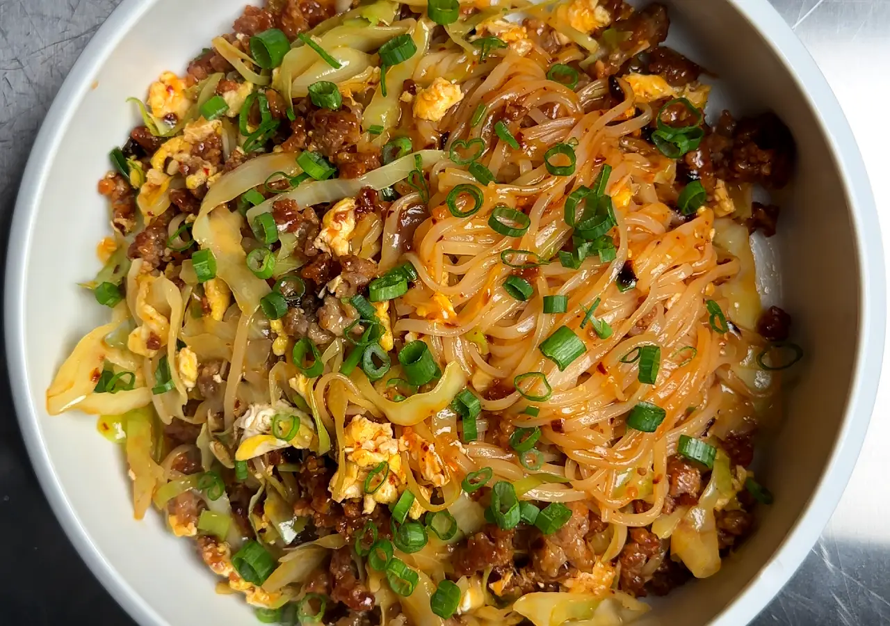 Chili Crispy Pork Noodles: wide rice noodles in glossy chili oil sauce with crumbled ground pork, sliced cabbage, scrambled egg, and scallions in a white bowl